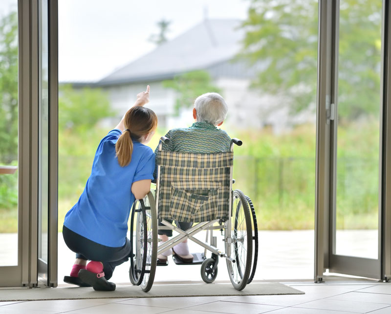 Junge weibliche Pflegekraft zeigt einer alten Person im Rollstuhl die Natur durch eine Glastür.